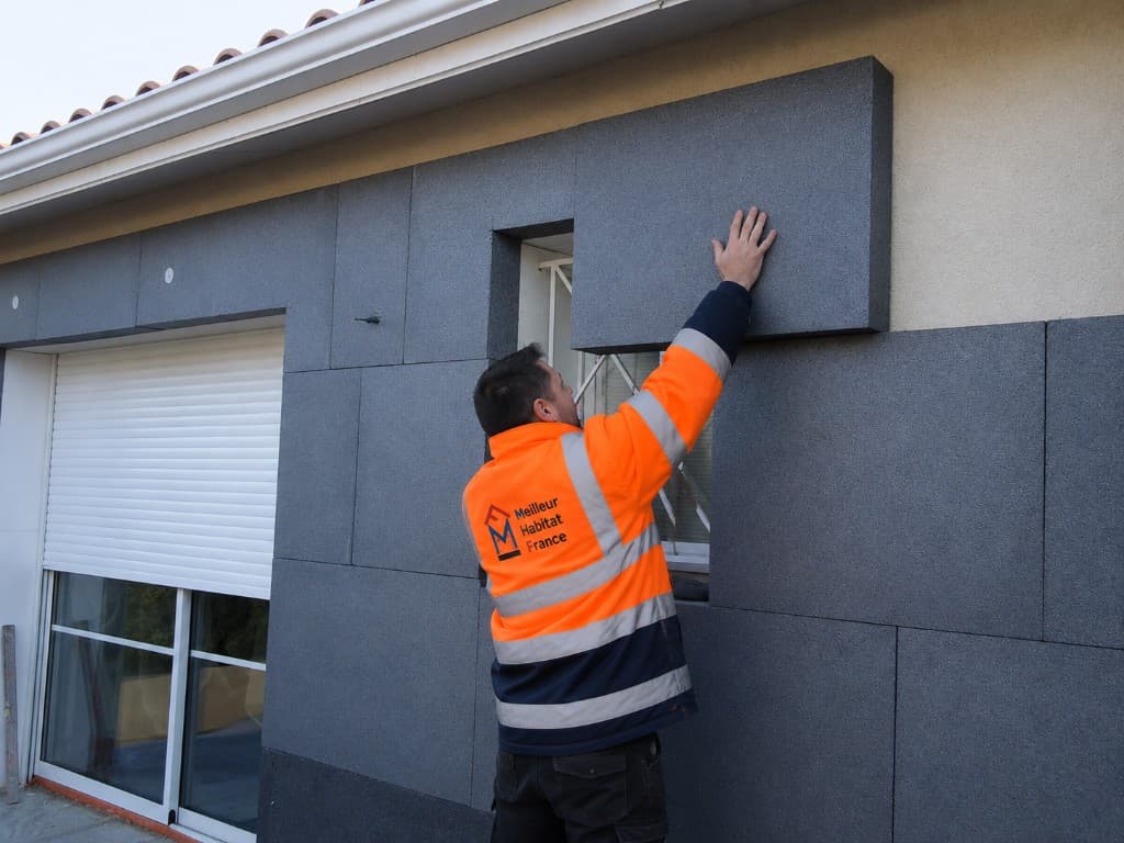 Artisan MHF en tenue haute-visibilité posant des panneaux d'isolation thermique par l'extérieur (ITE) sur la façade d'une maison individuelle.